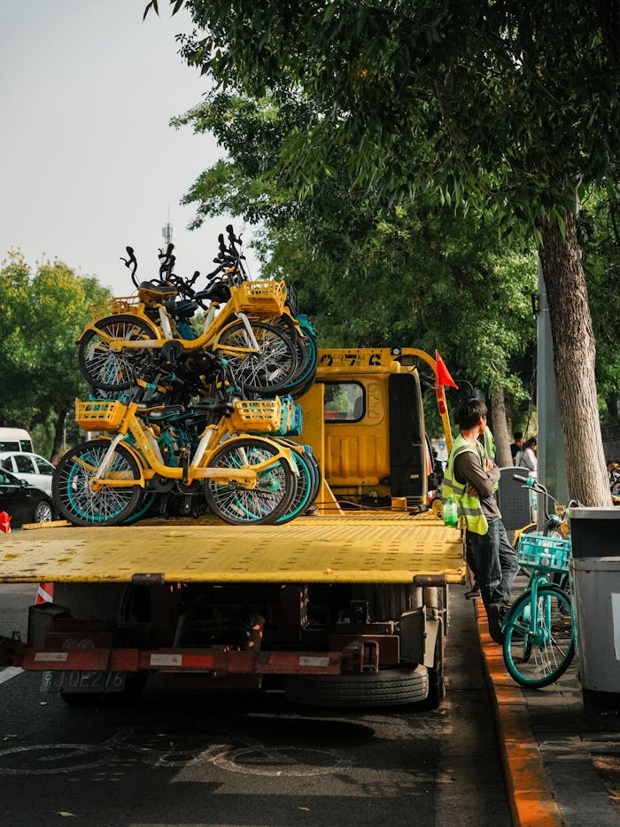 Urban scene with a tow truck carrying multiple yellow rental bikes on a tree-lined street.